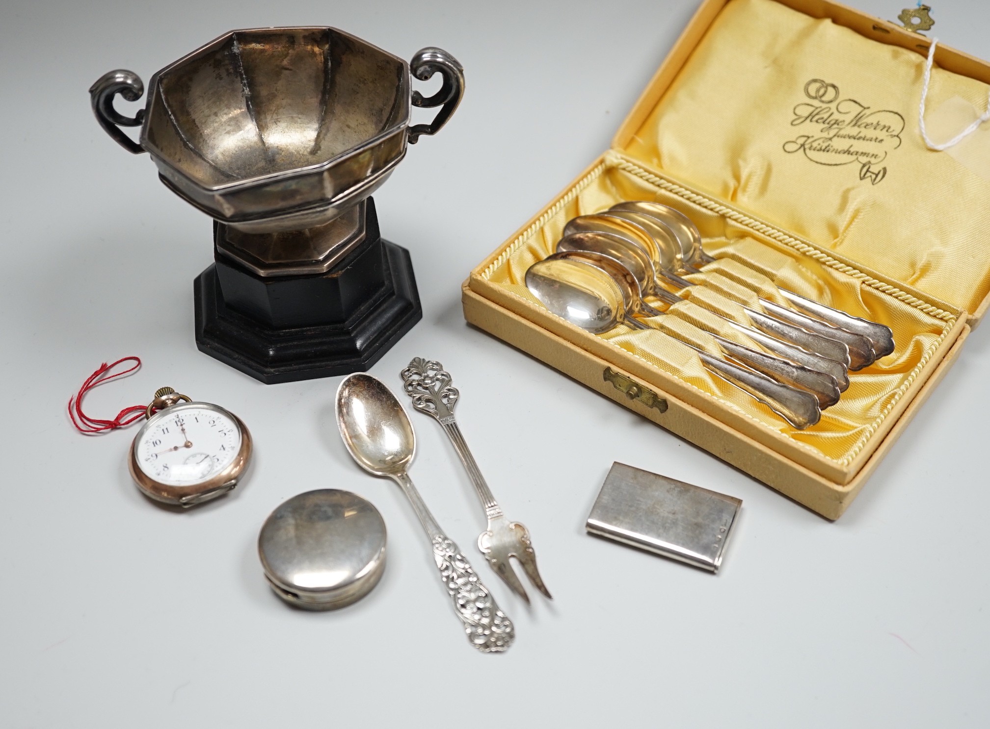 A late Victorian silver vesta case, a silver pill box, a small silver trophy cup, a cased set of six Swedish white metal coffee spoons, a similar spoon and fork and an early 20th century tow colour white metal fob watch.